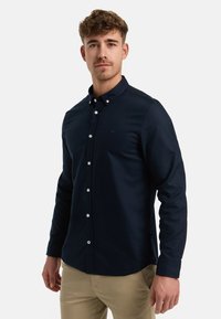 Nuori mies kiharat hiukset, yllään laivastonsininen nappipaita ja beiget housut, seisoo valkoista taustaa vasten.