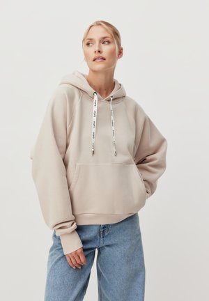 Donna che indossa una felpa beige e jeans blu chiaro, in piedi con una mano in tasca contro uno sfondo bianco uniforme.