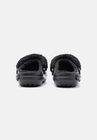 Črne klompice z mehko podlogo, odprto zadaj, teksturirano podplatom in pisanimi gumbastimi detajli na peti. Mehko gradivo in zaobljen oblik.