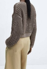 Pull en maille marron présentant un motif ajouré ample et une bordure côtelée, associé à un pantalon beige clair avec poches arrière.