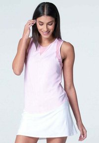 Mujer con cabello oscuro y lacio, vistiendo una blusa sin mangas de color rosa claro y una falda blanca, mirando hacia abajo y tocándose el cabello, contra un fondo gris liso.