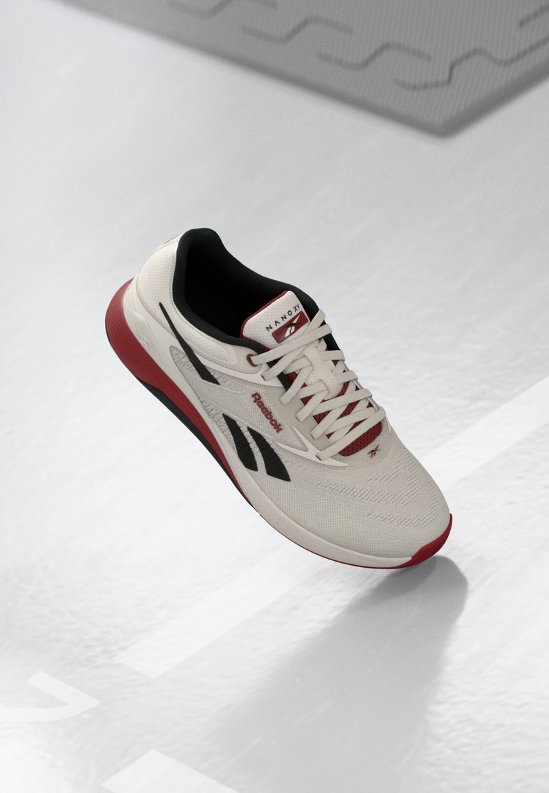 Hvit sportssko med teksturert overdel, svarte detaljer, rød såle og røde snøringer. Har Reebok-merke og et strømlinjeformet design.