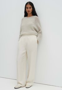 Donna con capelli lunghi e scuri che indossa un maglione beige lavorato a maglia, pantaloni ampi color crema e scarpe color crema, in piedi contro una parete bianca.