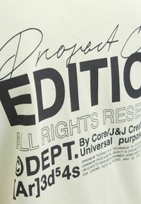 Camiseta de color beige claro con texto negro en negrita, que incluye "EDICIÓN DEL PROYECTO" y "DEPTO", con diferentes estilos y tamaños de fuente. Impresión gráfica texturizada.