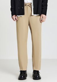 Khaki-Hosen mit entspannter Passform, elastischem Kordelzugbund und glatter Textur. Kombiniert mit schwarzen Schuhen und einem dunklen Blazer.