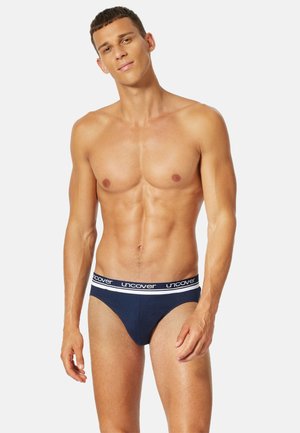 Junger Mann mit kurzen Haaren, der marineblaue Unterwäsche mit weißem "uncover"-Bund trägt, vor einem schlichten hellen Hintergrund stehend.