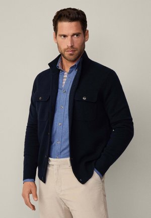 Navy cardigan with a high collar, made of knit material, featuring two chest pockets. Worn over a light blue checked shirt and beige pants.
