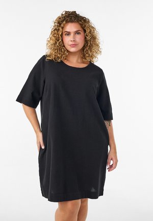 Žena s kučeravými blond vlasmi, ktorá nosí voľné čierne šaty s krátkym rukávom, stojí na bielom pozadí.