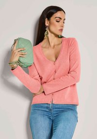 Roze cardigan met een V-hals, knopen aan de voorkant en lange mouwen. Een paar druppelvormige oorbellen en een mintgroene gewatteerde handtas met een kettingstrap.
