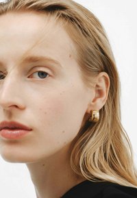 Pendientes de aro dorados con un diseño apilado y estriado. Textura metálica suave, de tamaño mediano, llevados por una persona de piel clara y cabello liso.