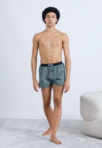 Zöld kockás anyagból készült boxeralsó fekete derékpánttal, amelyen a "BOSS" márkajelzés látható. A modell világos háttér előtt áll.