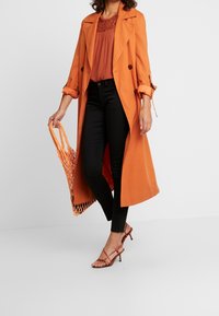 Trench coat largo naranja con mangas enrolladas sobre una camiseta de color óxido, combinado con jeans skinny negros y tacones rojos con tiras, llevando una bolsa de malla.