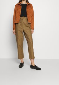 Giacca in suede marrone, top nero, pantaloni a vita alta color nocciola e mocassini neri. La giacca ha bottoni metallici; i pantaloni presentano una cintura visibile.