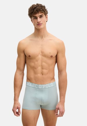 Homme torse nu aux cheveux bouclés portant un boxer gris clair avec une ceinture portant l'inscription "MARC O'POLO", debout devant un fond uni.