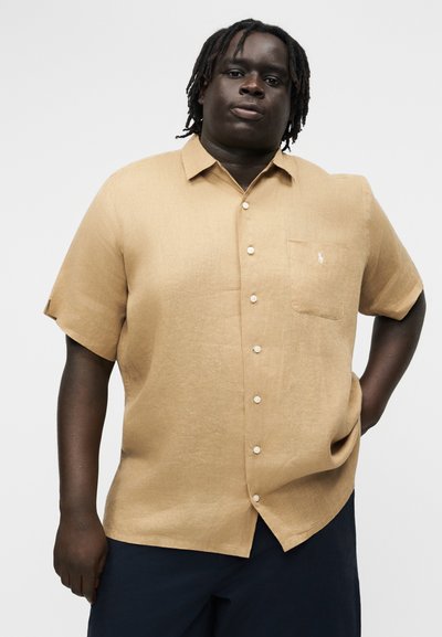 Mees tumeda nahaga ja dreadlockidega, kellel on seljas beež lühikeste varrukatega nööpidega särk ja tumedad püksid, seisab lihtsa tausta ees.