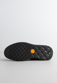 Czarna gumowa podeszwa buta z teksturowanym heksagonalnym wzorem, z pomarańczowym okrągłym logo i małą zieloną kropką po boku.