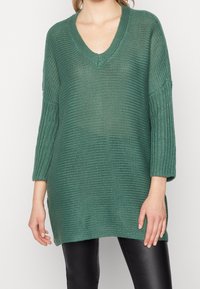 Grön, stickad oversized tröja med djup v-ringning, ribbade ärmar och sidslitsar, tillsammans med svarta byxor. Texturerad, lös passform.