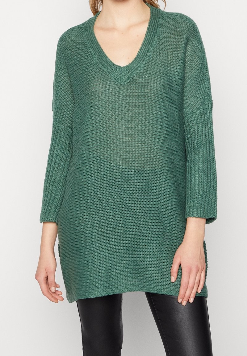 Grön, stickad oversized tröja med djup v-ringning, ribbade ärmar och sidslitsar, tillsammans med svarta byxor. Texturerad, lös passform.
