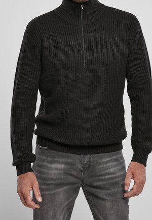 Homme portant un pull en maille noir à demi-fermeture éclair et un jean noir délavé, debout avec les mains détendues le long du corps sur un fond uni.