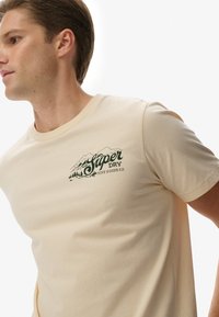 Camiseta de algodón beige con un gráfico de montaña verde oscuro y texto que dice "Super Dry Fine Goods Co." Diseño de cuello redondo y mangas cortas.
