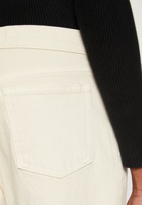 Denim de couleur crème avec une poche arrière, associé à un pull noir à côtes. La texture du tissu est visible, mettant en avant le contraste des matériaux.