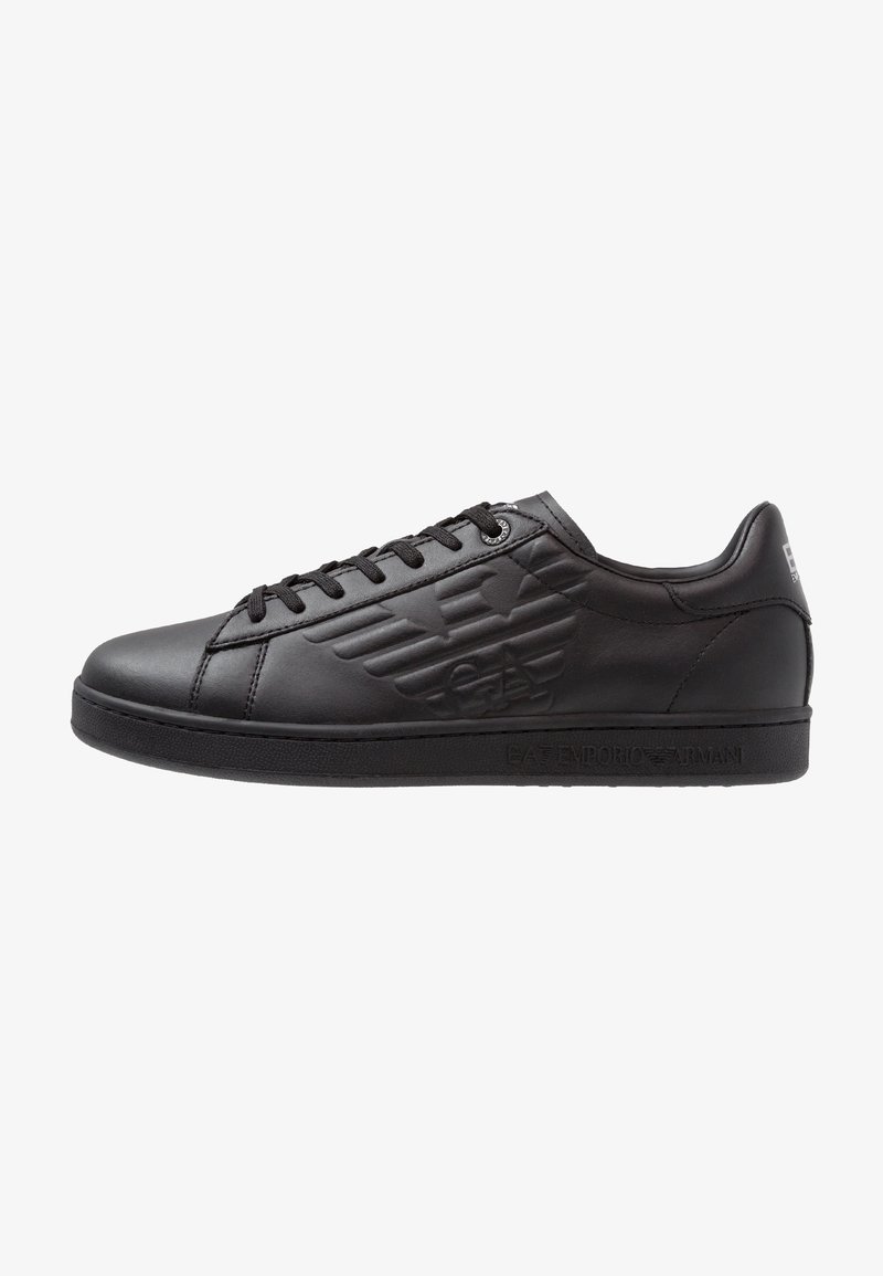 Baskets basses en cuir noir avec logo embossé sur le côté, lacets noirs et semelle plate, présentées en profil sur fond blanc.