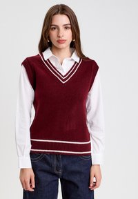 Gilet tricoté bourgogne avec des bords blancs, design en V, porté sur une chemise blanche à col, associé à un jean bleu foncé. Texture douce.