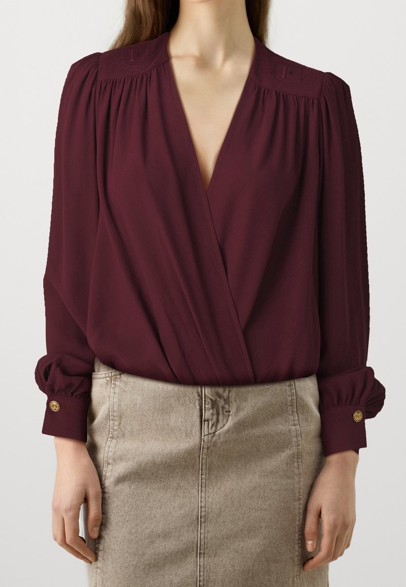 Blusa avvolgente bordeaux con scollo a V profondo, spalle arricciate e maniche a palloncino, con dettagli di bottoni sui polsini; abbinata a una gonna beige.