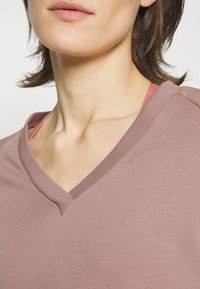 Top a scollo a V in morbido tessuto mauve, con colletto a coste e sottogiacca rosa sovrapposta visibile alle spalle.