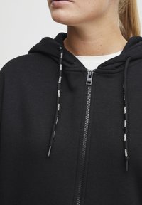 Hoodie zippé noir avec capuche, en tissu texturé, cordons de serrage avec logo de la marque, et fermeture éclair à l'avant.