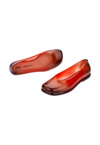 Zapatos planos transparentes rojos con una punta cuadrada redondeada, textura suave y brillante, y un branding minimalista en la suela interior.