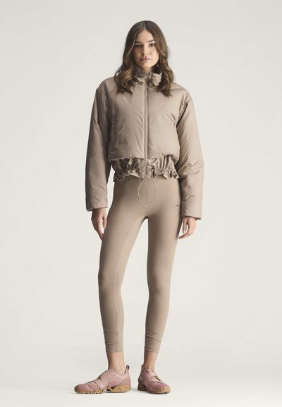 Frau trägt eine beige kurze Steppjacke, passende hoch taillierte Leggings und rosa Sportschuhe und steht vor einem einfarbigen Hintergrund.