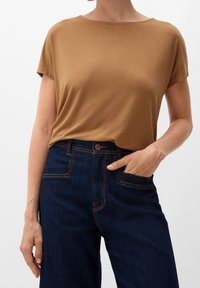 Brun kortærmet top lavet af blødt stof, parret med mørkeblå høj talje jeans med orange syninger og sidelommer.