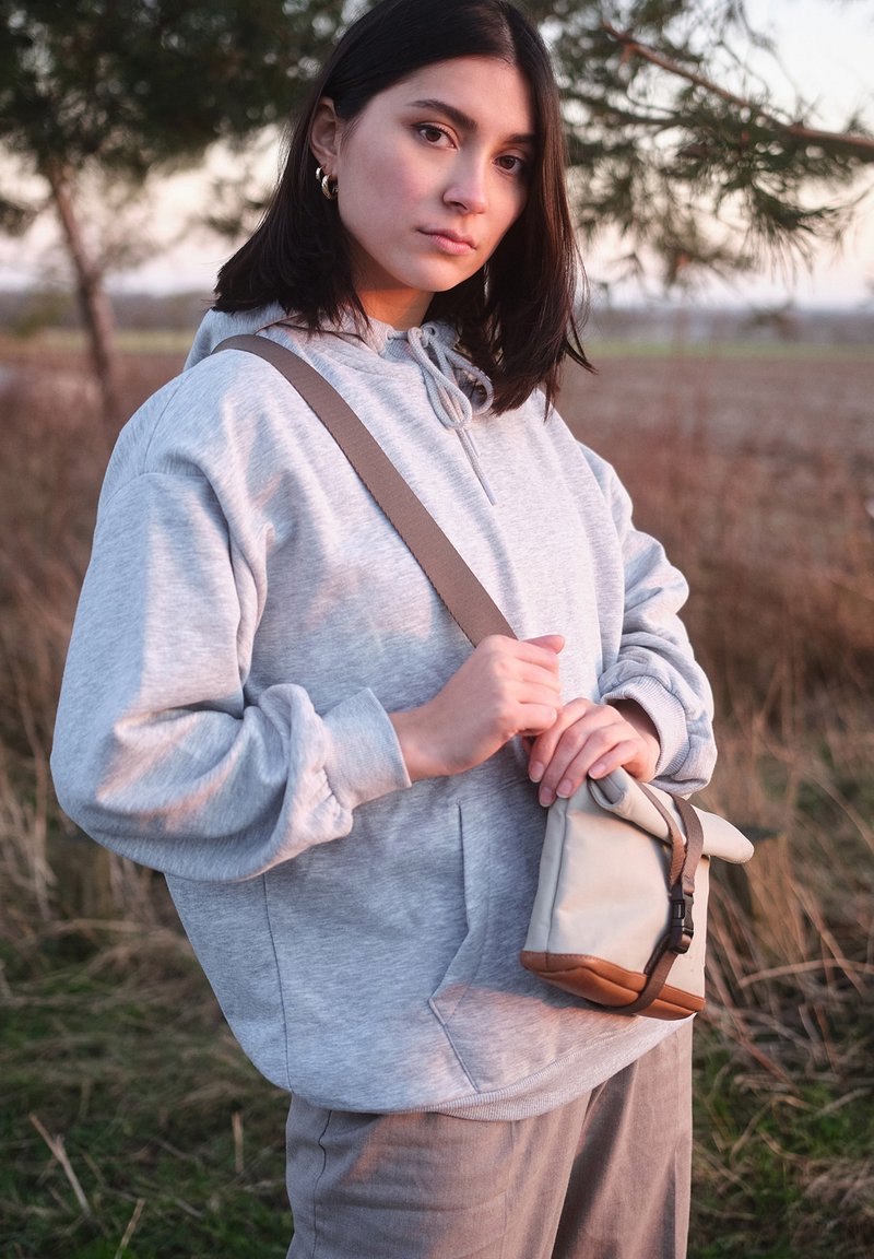 Felpa grigia con cappuccio e tasca frontale, abbinata a una borsa a tracolla beige con fondo marrone e tracolla regolabile. Sfondo di un ambiente naturale.