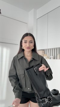 Schwarze Lederstiefel bis zum Knie mit metallischem Schnallendetail, glatter Textur und flacher Sohle. Das Model hält einen Stiefel in der Hand und trägt eine olivgrüne Jacke.