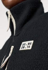Must on must fleece-tagi, mille on kõrge krae, elevandiluust tõmblukk ja ristkülikukujuline plaastriga, millel on mustal kreemil numbrid 55, 95, 09 ja 14.