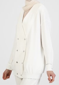 Witte, geribbelde lange jas met een dubbelgeknoopt ontwerp, voorzien van metalen drukknopen en een getailleerde tailleband. Lichtgewicht stof.