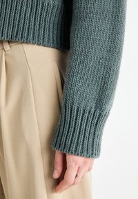 Gros plan sur une main avec un petit tatouage de croix sur le doigt, vêtue d'un pull en maille épaisse couleur bleu sarcelle et d'un pantalon plissé beige.