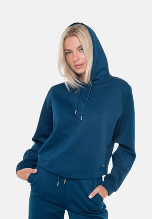 Donna con capelli biondi che indossa una felpa con cappuccio blu e pantaloni abbinati, con le mani nelle tasche, su uno sfondo semplice.