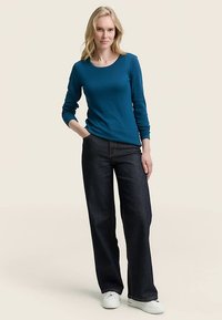 Turkizna majica z dolgimi rokavi, kombinirana s temno modrimi širokimi kavbojkami in belimi supergami. Tkanina je videti gladka in udobna.