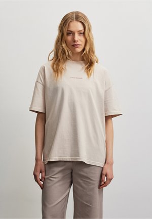Młoda kobieta z falowanymi blond włosami, ubrana w luźny, jasnobeżowy T-shirt i spodnie w kolorze taupe na jednolitym szarym tle.