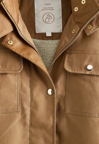 Veste marron résistante à la douche avec fermeture éclair à l'avant, boutons-pression, deux poches à rabat et doublure intérieure douce et texturée visible près du col.