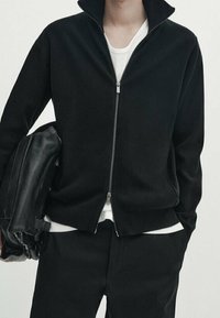 Giacca in pile nera con colletto alto e zip frontale, abbinata a una maglietta bianca. Il modello tiene una borsa in pelle nera.