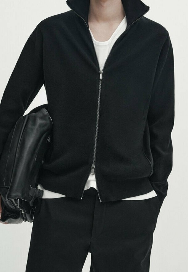Giacca in pile nera con colletto alto e zip frontale, abbinata a una maglietta bianca. Il modello tiene una borsa in pelle nera.