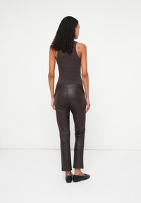 Top fără mâneci de culoare maro închis și pantaloni din piele maro, ajustați, care au o textură netedă și un design subțire. Pantofii negri cu talpă joasă completează ținuta.