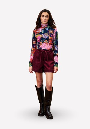 Vrouw in een kleurrijke, gestreepte blouse met lange mouwen, bordeauxrode hoge shorts met een gestrikte riem, en zwarte kniehoge laarzen, staand met het gezicht naar voren.