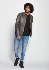 Graue Lederjacke mit Reißverschluss vorne, zwei Brusttaschen und glatter Textur, kombiniert mit schwarzem Shirt, blauen Jeans und schwarzen Stiefeln.