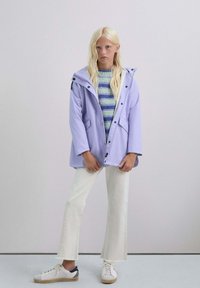 Chaqueta con capucha de color lavanda claro con botones de presión, dos bolsillos delanteros, llevada sobre un suéter a rayas azul y verde, combinado con vaqueros blancos acampanados y zapatillas deportivas.
