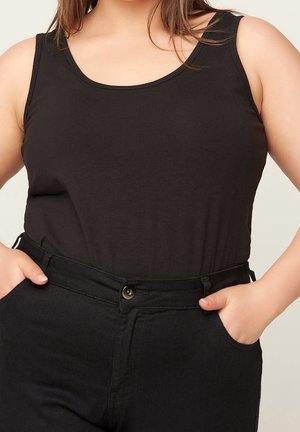 Vrouw draagt een mouwloze zwarte top die in een zwarte broek met hoge taille is gestopt, met handen in de zakken, tegen een neutrale achtergrond.