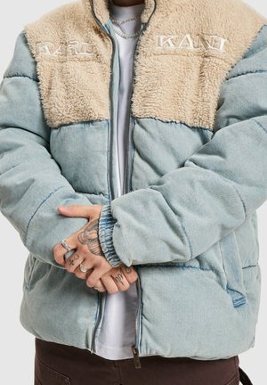 Lichtblauwe donsjas met beige fleece borstpanelen geborduurd met "KARL KANI," gedragen over een wit shirt, handen gekruist met zichtbare tatoeages en ringen.